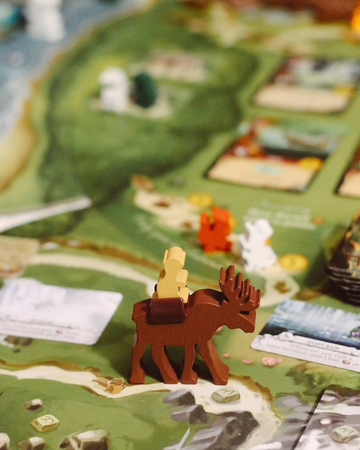 Close-up of the Everdell board with a brown wooden moose figure carrying a pale worker meeple across a green forest landscape dotted with resource tokens and cards.