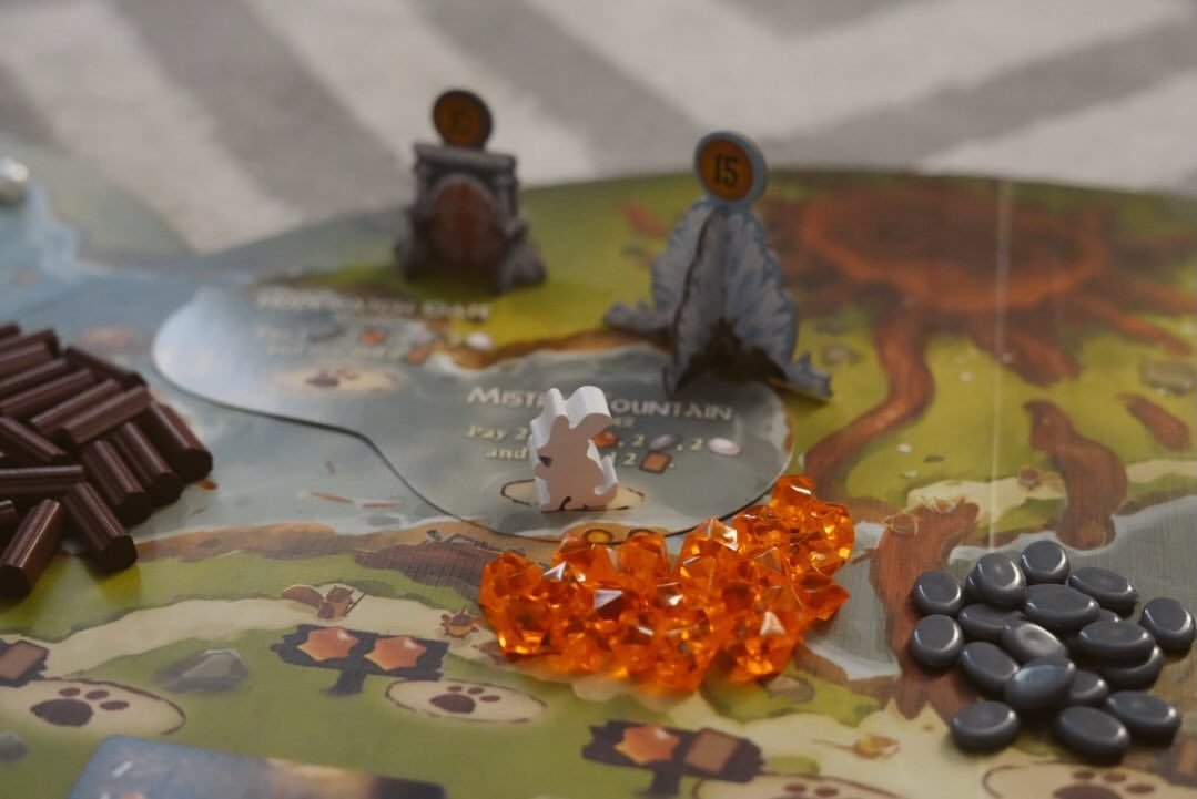 Macro view of Everdell components: a white rabbit worker meeple beside cardboard mountain standees, with piles of brown wood, orange gem and grey stone resource tokens.
