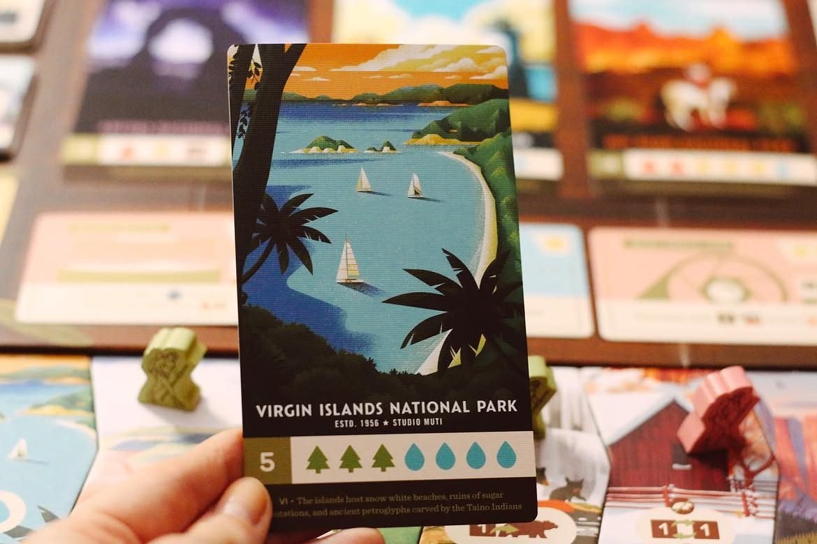 A hand holds the Parks card 'Virgin Islands National Park', a stylized bay with sailboats and palm trees, above the blurred game board.