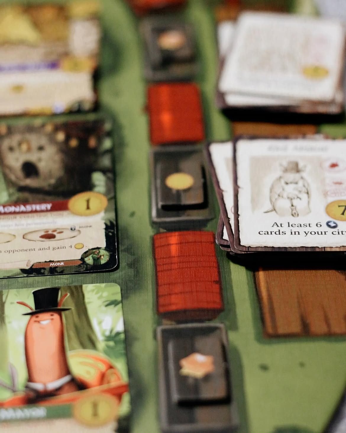 Close-up of Everdell cards, including a 'Monastery' card and a top-hatted critter 'Mayor' card, with stacked red gem tokens and a scoring card.