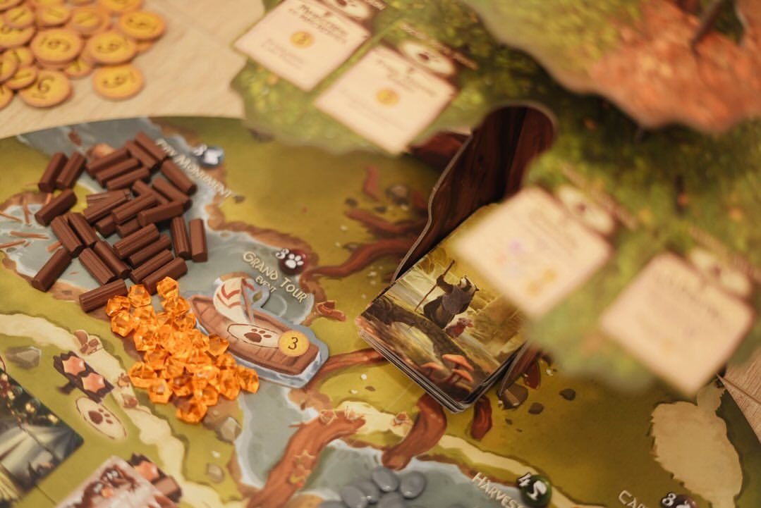 Warm close-up of the Everdell board with a 'Grand Tour' boat space, piles of brown wood and orange gem resources, gold point tokens and a card deck.